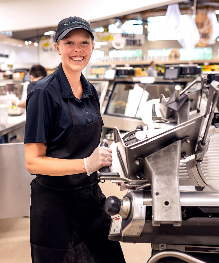a woman in a black hat and apron standing in a grocery store deli
